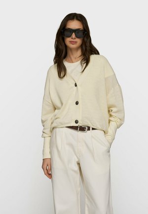 Kobieta w dużych czarnych okularach przeciwsłonecznych, kremowym rozpinanym kardiganie, dopasowanych spodniach i brązowym pasku, stojąca z jedną ręką w kieszeni.