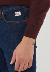 Jeans in denim blu con rivetti di rame, dotati di un'etichetta bianca con testo rosso sulla tasca anteriore, abbinati a un maglione bordeaux.