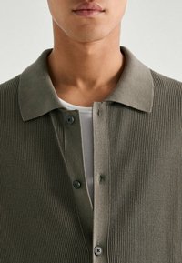 Olive green, ribbed knit cardigan with a fold-over collar, button closure, and two black buttons visible at the front. White undershirt beneath.
