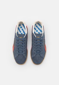 Modré semišové tenisky s červenými akcenty a gumovými podrážkami. Mezi jejich vlastnosti patří perforované špičky a potištěné stélky se znakem "Reebok".