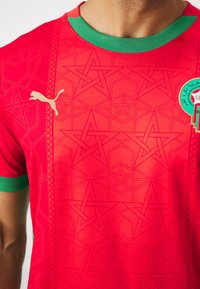 Maglia sportiva rossa con un motivo a stella geometrica, colletto verde e logo Puma dorato. Presenta un tessuto traspirante e testurizzato.