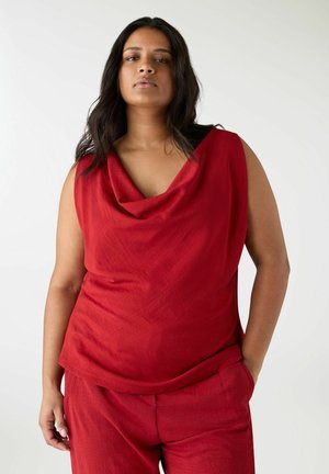 Donna con capelli lunghi e scuri che indossa un top rosso senza maniche e pantaloni rossi abbinati, in piedi con una mano in tasca contro uno sfondo bianco.