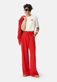 Witte T-shirt met rode lippenafbeelding, rode hooggetailleerde broek, een rode blazer over de schouders gedrapeerd en slip-on schoenen met luipaardprint. Zonnebril als accessoire.