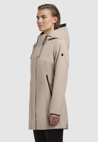 Veste imperméable beige avec capuche, dotée de poches zippées noires et de cordons de serrage, texture lisse et design mi-long.