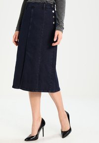 Denimová sukně tmavě modré barvy, po kolena, se rovný střihem a knoflíkovou aplikací na boku, nošená s černými vysokými podpatky.