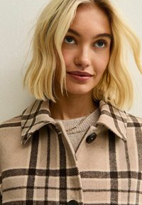 Blond kvinna med axellångt hår, iklädd en beige rutig kappa och guldkedja, som tittar något uppåt och ler mjukt.