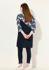 Mörkblå klänning med mönstrade blå och krämfärgade ärmar, i kombination med mörkblå leggings med spetsdetaljer vid fållen och lätta, avslappnade skor.