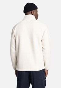 Crème fleecejas met een hoge kraag en lange mouwen, met een gestructureerd oppervlak. Gedragen met donkere broeken en een marineblauwe baret.