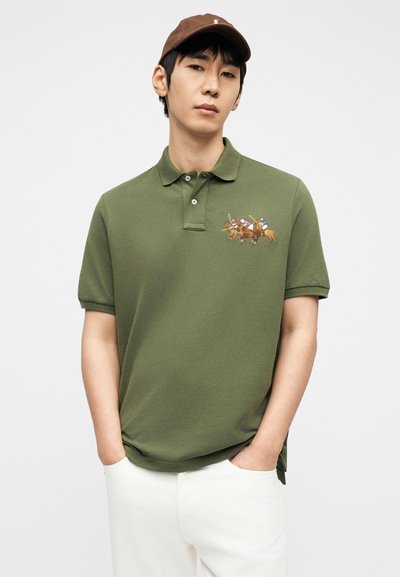 Άνδρας που φοράει καφέ καπέλο, χακί πράσινο πόλο πουκάμισο με κεντημένους παίκτες πόλο και λευκό παντελόνι, στέκεται με τα χέρια στις τσέπες.