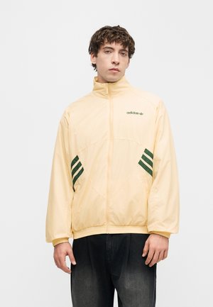 Młody mężczyzna noszący jasnożółtą kurtkę Adidas na zamek błyskawiczny z ciemnozielonymi paskami i logo, połączoną z ciemnymi spodniami, na jednolitym tle.