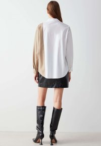 Camicia a maniche lunghe con parte posteriore bianca, lato sinistro beige e una mini gonna in pelle nera. Stivali alti fino al ginocchio neri e lucidi con tacco a stiletto.