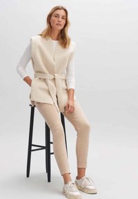 Beige Fleece-Weste mit Wickeldesign und Gürtel, über einem weißen Langarmshirt getragen. Kombiniert mit hellbeigen Slim-Pants und Sneakern.