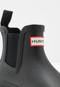 Stivale in gomma nera alla caviglia con pannelli laterali elasticizzati, un anello in tessuto sul retro e un'etichetta con "HUNTER" in bianco su sfondo rosso.