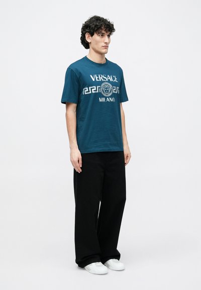 Zaļa kokvilnas T-krekls ar baltu "VERSACE MILANO" logotipu un grieķu atslēgas rakstu; kopā ar melnās platajās biksēs un baltām sporta kurpēm.