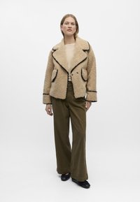 Beige Kunstfelljacke mit schwarzem Besatz, breiten Revers und zwei vorderen Taschen, kombiniert mit olivgrünen weit geschnittenen Hosen und schwarzen Schuhen.