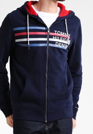 Mann trägt marineblauen Kapuzenpullover mit Reißverschluss, rotem Kapuzeninnenfutter, weißen Kordeln, horizontalen roten, weißen und blauen Streifen und dem Schriftzug „Tommy Hilfiger Denim“.