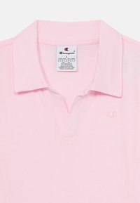 Ljust rosa kortärmad polo med krage, som har en liten broderad logotyp och en etikett som visar storlek och skötselråd.