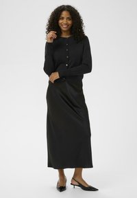 Cárdigan negro con cuello redondo, cierre de botones, llevado sobre una falda larga de satén negra. Completado con tacones negros de punta. Textura suave.