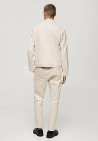 Light beige overshirt and matching trousers made of textured fabric. Features double back pockets, clean lines, and a simple design. Black footwear.