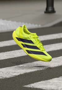 Chaussure de course jaune néon avec des accents violets, design léger, forme effilée et semelle texturée, suspendue au-dessus d'un passage piéton.