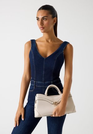 Beige leren handtas met een gebogen ontwerp, gouden hardware en twee handvatten; gedragen bij een donkerdenim jumpsuit met een V-hals.
