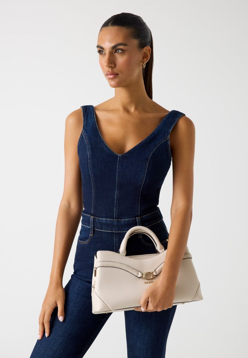 Beige leren handtas met een gebogen ontwerp, gouden hardware en twee handvatten; gedragen bij een donkerdenim jumpsuit met een V-hals.