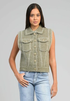 Femme aux longs cheveux foncés portant un gilet vert sans manches à boutons avec poches avant et un jean bleu clair, sur fond gris uni.
