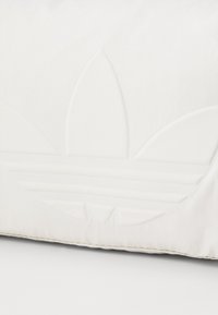 Cojín blanco con diseño de logo en relieve que presenta formas de hojas y líneas horizontales. El material parece suave y mate.
