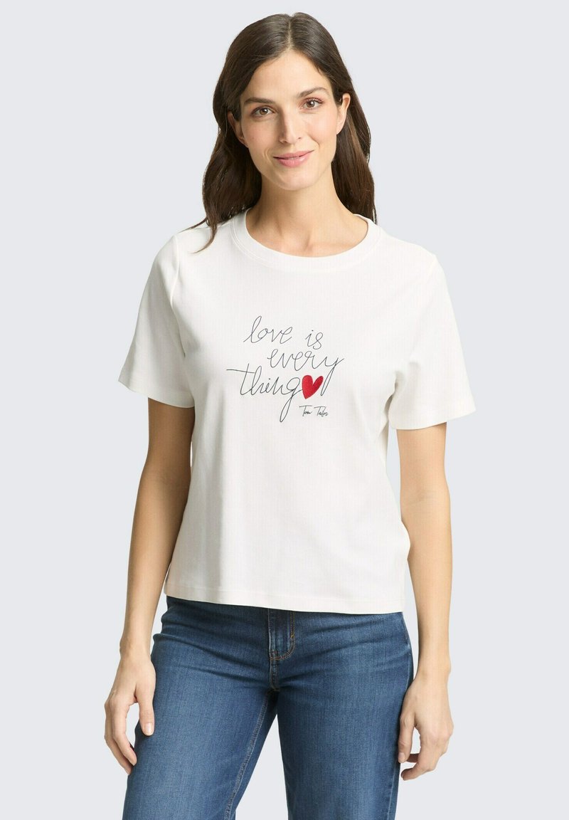 Femme aux longs cheveux bruns portant un t-shirt blanc avec le texte "love is everything" et un cœur rouge, associée à un jean bleu sur un fond uni.