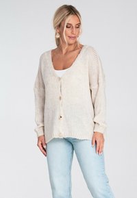 Jasnobeżowy sweter z dzianiny z dekoltem w serek, pięcioma guzikami i prążkowanymi mankietami. Nosi się go na białej bluzce w połączeniu z jasnoniebieskimi dżinsami.