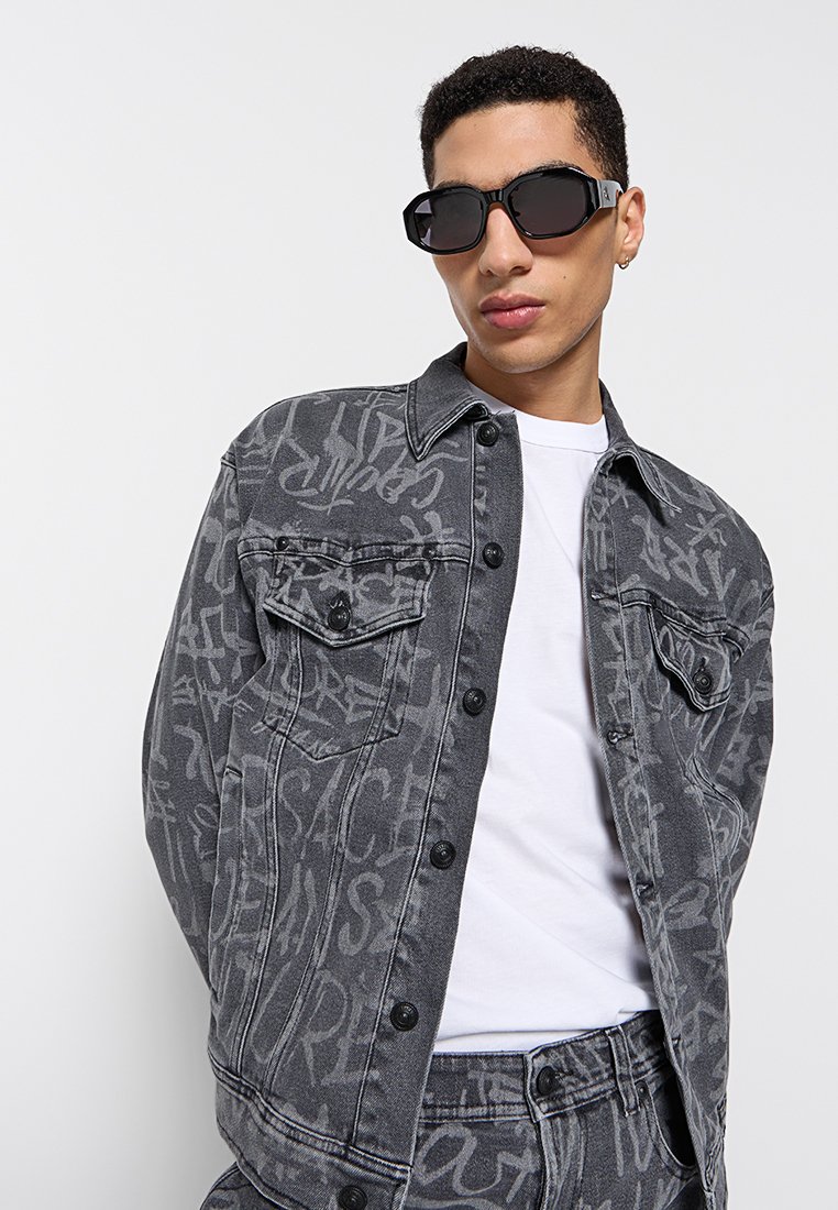 Chaqueta de mezclilla negra con patrones en estilo graffiti en gris, dos bolsillos en el pecho y cierre de botones, combinada con una camiseta blanca y gafas de sol negras.