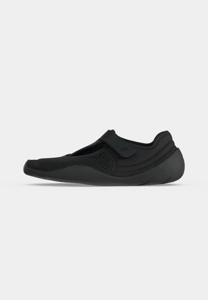 Zapato negro tipo slip-on de malla con cinta elástica sobre el empeine y suela de goma redondeada, diseñado para comodidad y flexibilidad.