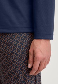 Navy blauwe langemouwen top met een gladde textuur, gecombineerd met gestreepte leggings met een oranje geometrisch ontwerp tegen een donkere achtergrond.
