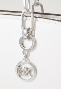 Hopeinen Michael Kors -logo-riipus roikkuu valkoisesta teksturoidusta käsilaukusta, jossa on ruskeat ompeleet.