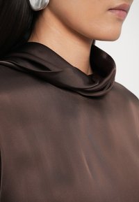 Καφέ σατέν τοπ με λαιμόκοψη σε σχήμα κάλυκα, που διαθέτει λεία υφή. Συνοδεύεται από μεγάλο μεταλλικό σκουλαρίκι.