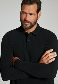 Schwarzes Langarm-Poloshirt mit Kragen, aus strukturiertem Stoff und mit einer Knopfleiste mit drei Knöpfen. Das Model hat die Arme in neutraler Pose verschränkt.