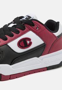 Zapatilla negra, blanca y marrón con un upper de cuero sintético texturizado. Presenta un logotipo bordado prominente y cordones negros planos.
