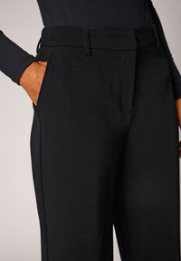 Los pantalones negros de talle alto presentan un corte entallado, dos bolsillos laterales y una textura de tela suave. La cintura tiene trabillas para cinturón para su ajuste.