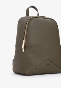 Zaino in pelle verde oliva con forma strutturata, accessori in metallo dorato, superficie testurizzata e un dettaglio di design triangolare alla base.