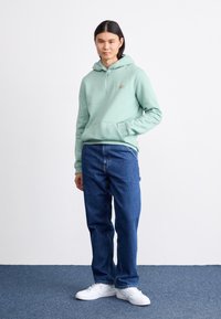 Light green hoodie with a front pocket and adjustable hood, paired with dark blue jeans and white sneakers, standing on a blue carpet.