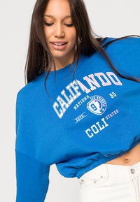 Sudadera azul con texto gráfico en blanco y rosa "CALIFANDO" y logo. Presenta un corte holgado, cuello redondeado y dobladillo acanalado.