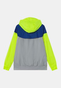 Giacca a vento con cappuccio caratterizzata da un corpo grigio, maniche giallo neon e accenti blu navy sulle spalle. Texture liscia, polsini elasticizzati.