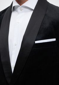 Άνδρας που φοράει μαύρο σακάκι τούξεντο με σατέν πέτα, λευκό πουκάμισο κουμπωμένο και λευκή μαντηλίτσα στην τσέπη του σακακιού.
