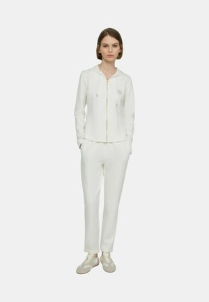 Giovane donna in piedi con le mani nelle tasche, indossa una felpa con cappuccio bianca con zip, pantaloni abbinati e scarpe da ginnastica di colore chiaro su sfondo uniforme.