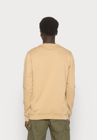 Sudadera de algodón beige con mangas largas, puños acanalados y cuello redondo. Presenta un pequeño parche con logo cerca del dobladillo.