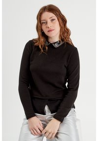 Black long-sleeve top featuring a contrasting black and white embellished collar, with a straight cut and textured fabric. Silver pants visible.