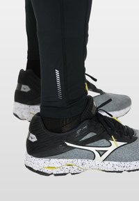 Zwarte sportlegging met reflecterende lijn details, gecombineerd met grijze hardloopschoenen met een gespikkelde witte zool en gele accenten.