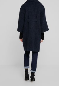 Granatowy płaszcz z mieszanki wełny o luźnym kroju, szerokich rękawach i detalem w postaci kieszeni z tyłu, noszony na dopasowanych dżinsach i botkach na obcasie.