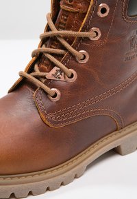 Botte en cuir marron avec bout rond, couture beige et œillets en métal. Équipée d'un système de laçage et d'une semelle en caoutchouc texturé pour une meilleure adhérence.