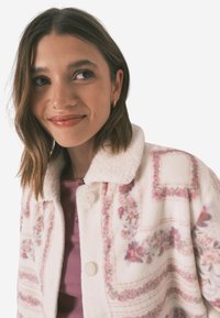 Fleecejacka med ett rosa blommönster på en ljus bakgrund, med en vit, fluffig krage och två knappstängningar.